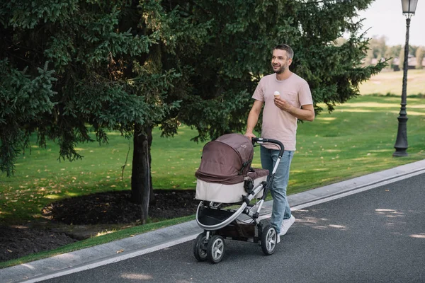 Smiling father walking with baby carriage and ice cream in park and looking away — Stock Photo