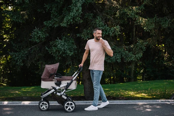 Smiling father walking with baby carriage and eating ice cream in park — Stock Photo