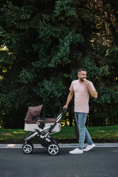 Усміхнений батько ходить з дитячою каретою і морозивом в парку і озирається назад — стокове фото