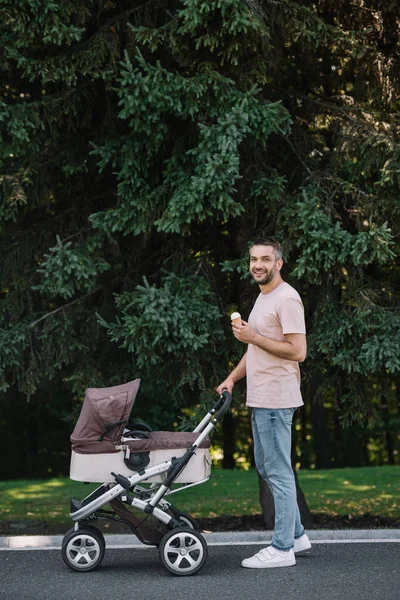 Vista lateral do pai sorrindo andando com carrinho de bebê e sorvete no parque — Fotografia de Stock