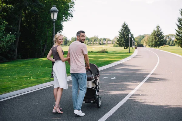 Vue arrière des parents marchant avec une voiture de bébé sur la route dans le parc et regardant la caméra — Photo de stock