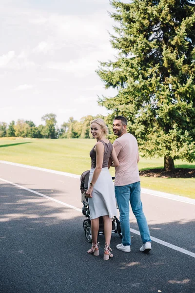 Parents walking with baby carriage on road in park and looking at camera — Stock Photo