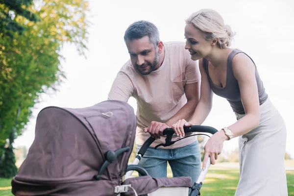 Cheerful parents looking at baby carriage in park — Stock Photo