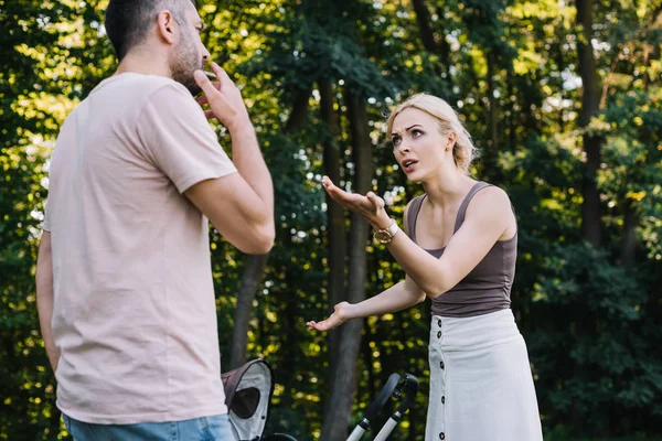 Père fumant la cigarette près de la voiture de bébé dans le parc, mère gesticulant et le regardant — Photo de stock