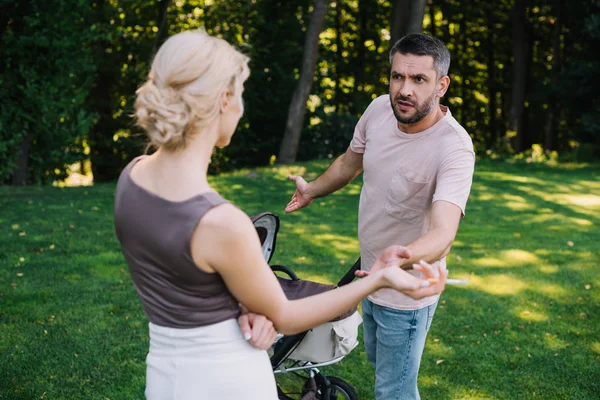 Mother smoking cigarette near baby carriage in park, angry father gesturing and looking at her — Stock Photo