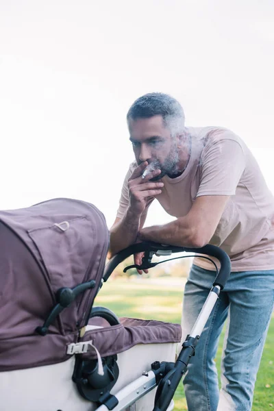 Padre fumar cigarrillo cerca de bebé cochecito en el parque - foto de stock
