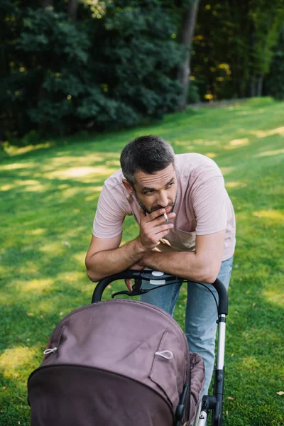 Father smoking cigarette near baby carriage in park and looking away — Stock Photo
