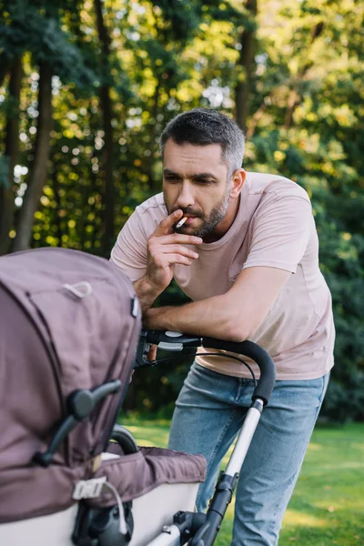 Father smoking cigarette near baby carriage in park — Stock Photo