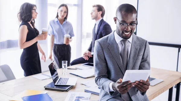 Enfoque selectivo de sonreír hombre de negocios afroamericano utilizando tableta y colegas en el lugar de trabajo en la oficina - foto de stock