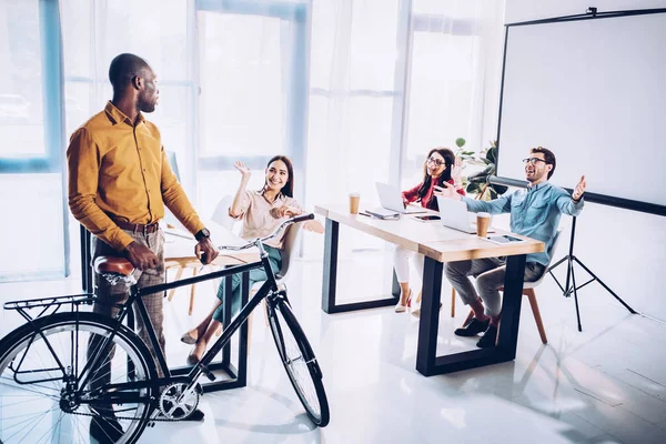 Multicultural business people greeting african american colleague with bicycle in office — Stock Photo