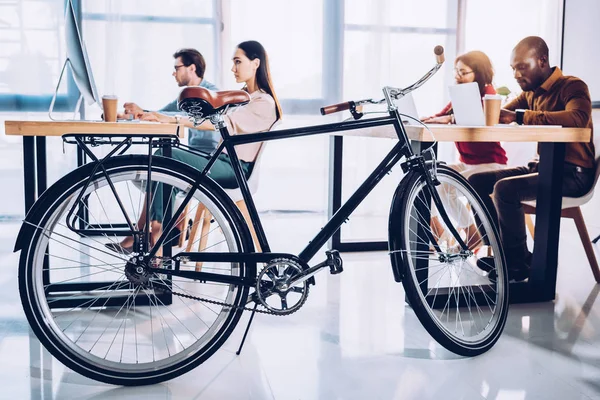 Side view of bicycle and multicultural business people working in office — Stock Photo