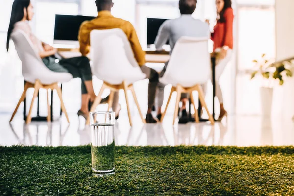 Selective focus of glass of water and multiethnic business people sitting at table in office — Stock Photo