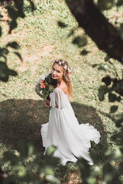 Blick aus der Vogelperspektive auf die schöne zarte junge Braut, die einen Blumenstrauß hält und im Freien in die Kamera lächelt — Stockfoto