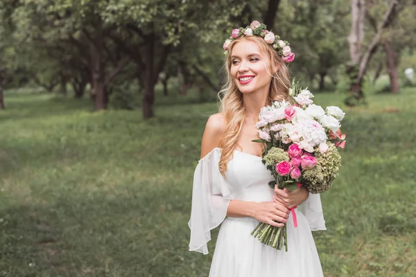 Bela noiva jovem feliz segurando buquê de flores e olhando para longe no parque — Fotografia de Stock