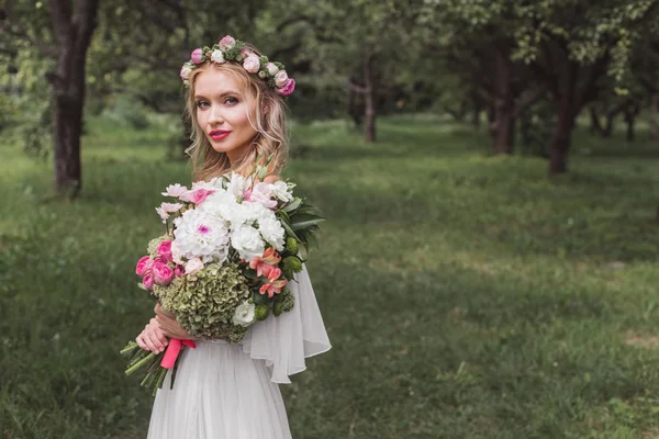 Novia en corona floral y vestido de novia con ramo de flores y mirando a la cámara al aire libre - foto de stock