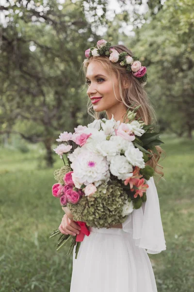 Красивая улыбающаяся молодая невеста, держащая свадебный букет и смотрящая в парк — стоковое фото