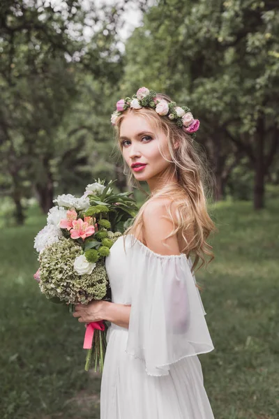 Zarte junge Braut in Blumenkranz und Brautkleid mit Blumenstrauß und Blick in die Kamera im Freien — Stockfoto