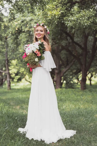 Bella felice giovane sposa tenendo bouquet da sposa e sorridendo alla macchina fotografica nel parco — Foto stock
