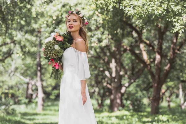 Beautiful smiling young bride in floral wreath holding bouquet of flowers and looking at camera — Stock Photo