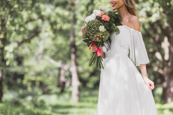 Schnappschuss einer lächelnden jungen Braut im Brautkleid mit Blumenstrauß im Freien — Stockfoto