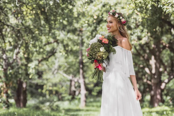 Schöne glückliche junge Braut hält Brautstrauß in der Hand und schaut im Park weg — Stockfoto