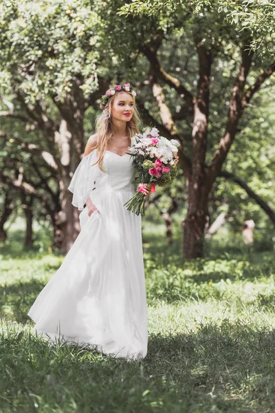 Belle jeune mariée heureuse en robe de mariée tenant bouquet de fleurs et marche dans le parc — Photo de stock