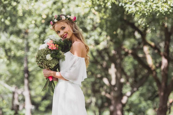 Hermosa joven novia en floral corona celebración ramo de flores y sonriendo a la cámara - foto de stock