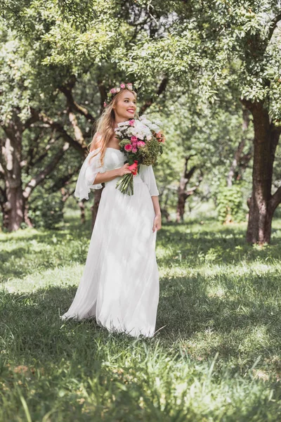 Schöne glückliche junge Braut hält Hochzeitsstrauß und geht im Park spazieren — Stockfoto