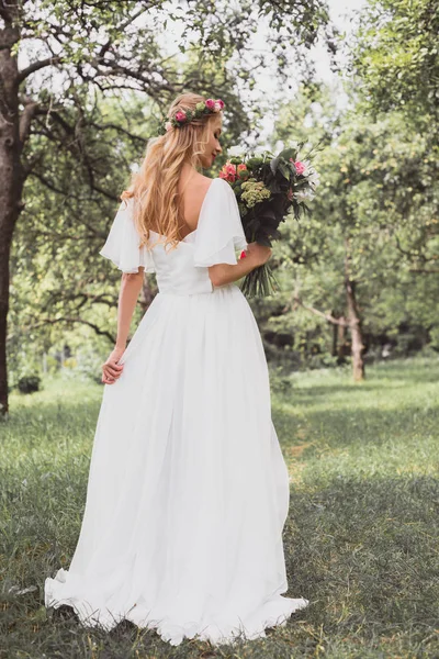 Vista posteriore della bella giovane sposa che tiene bouquet da sposa nel parco — Foto stock