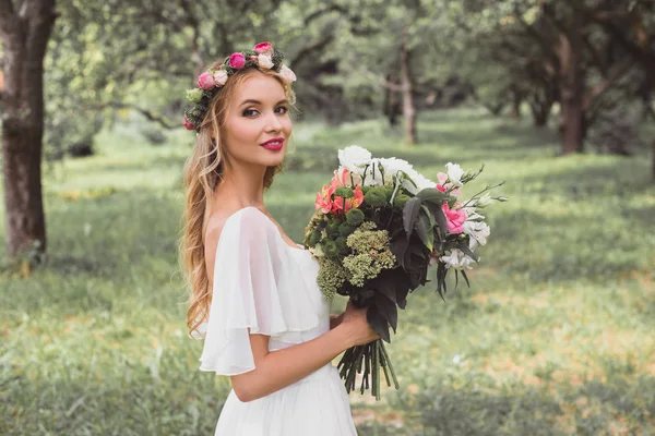 Mariée heureuse en robe de mariée et couronne florale tenant bouquet de fleurs et souriant à la caméra à l'extérieur — Photo de stock