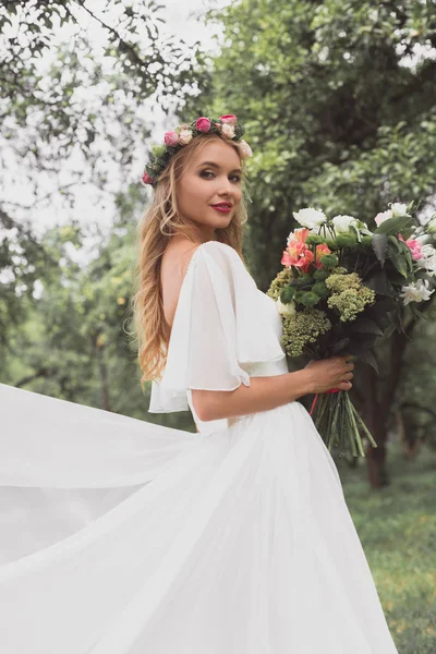 Noiva loira bonita em vestido de noiva e coroa floral segurando buquê de flores e sorrindo para a câmera ao ar livre — Fotografia de Stock