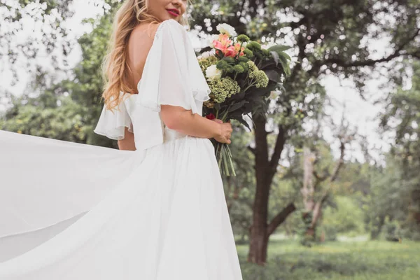 Cropped shot of young bride in wedding dress holding bouquet of flowers outdoors — Stock Photo