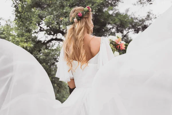 Back view of young blonde bride in wedding dress holding bouquet of flowers outdoors — Stock Photo