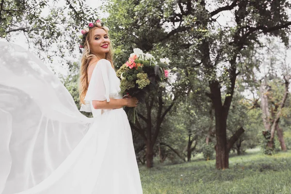 Happy blonde bride in wedding dress and floral wreath holding bouquet and smiling at camera in park — Stock Photo