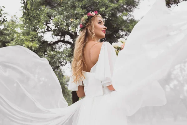 Vista de ángulo bajo de la novia rubia feliz en vestido de novia y corona floral mirando hacia otro lado en el parque - foto de stock
