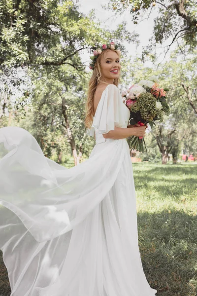 Bella giovane sposa in abito da sposa in possesso di bouquet di fiori e sorridente alla fotocamera nel parco — Foto stock
