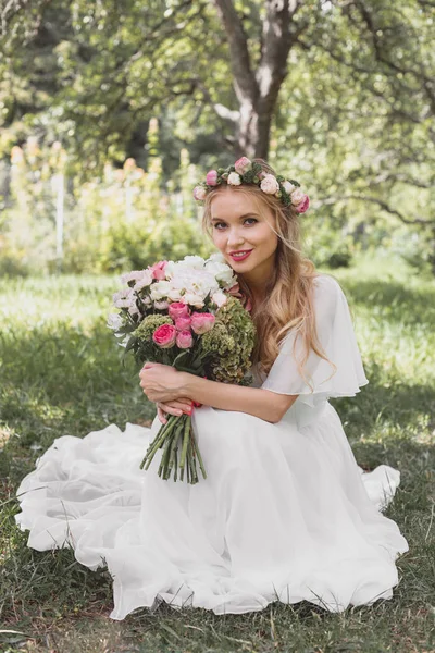 Atractiva novia joven celebración de ramo de bodas y sonriendo a la cámara en el parque - foto de stock