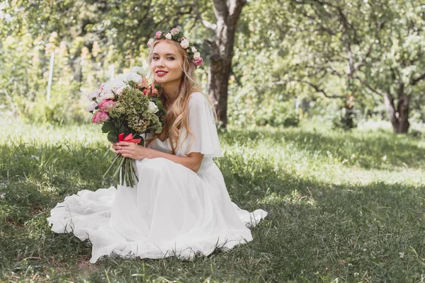 Beautiful young blonde bride holding wedding bouquet and smiling at camera in park — Stock Photo