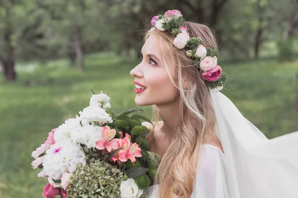 Side view of beautiful tender bride holding bouquet of flowers and looking up in park — Stock Photo