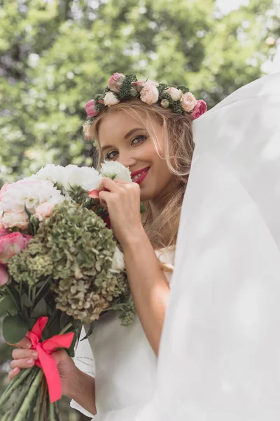 Low angle view of happy young bride in floral wreath and veil holding wedding bouquet and smiling at camera — Stock Photo