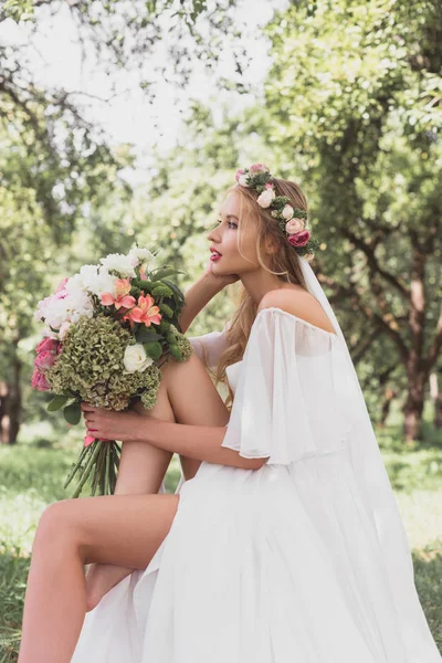 Вид сбоку на молодую застенчивую невесту, держащую свадебный букет и отводящую взгляд в парк — стоковое фото