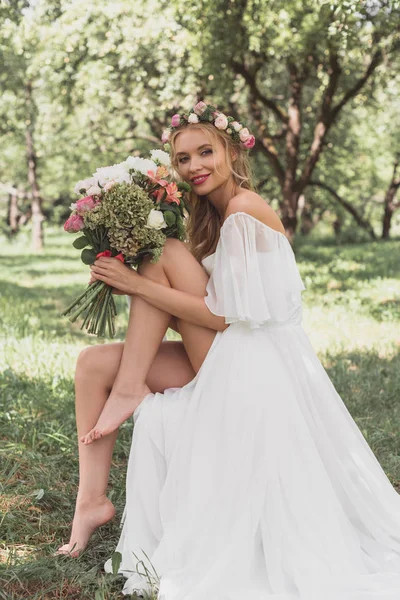 Belle jeune mariée pieds nus tenant bouquet de fleurs et souriant à la caméra — Photo de stock