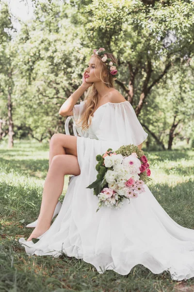 Schöne nachdenkliche junge Braut hält Brautstrauß und sitzt auf Stuhl im Park — Stockfoto