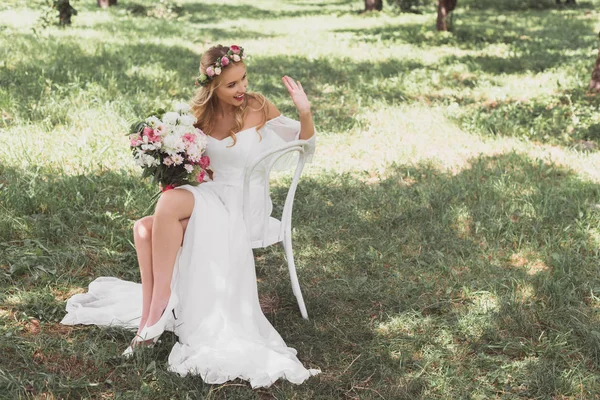 Vue grand angle de jeune mariée heureuse tenant bouquet de fleurs et agitant la main tout en étant assis sur la chaise à l'extérieur — Photo de stock