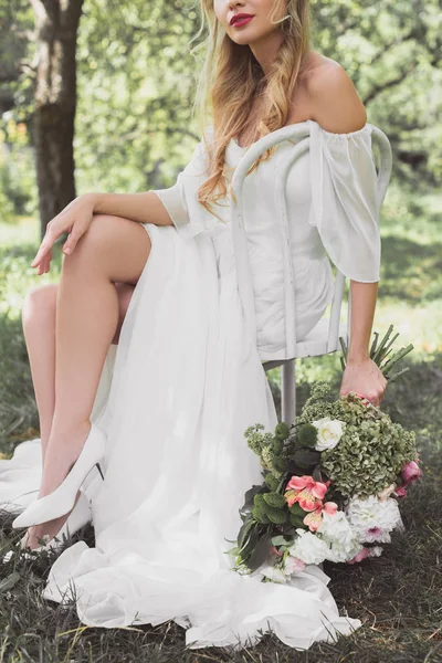 Schnappschuss der schönen blonden Braut mit Brautstrauß und im Freien auf einem Stuhl sitzend — Stockfoto
