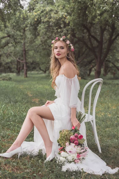 Hermosa joven novia con ramo de boda sentado en la silla y sonriendo a la cámara en el parque - foto de stock