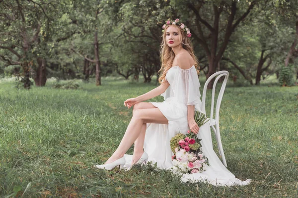 Beautiful young bride with wedding bouquet sitting on chair and looking at camera in park — Stock Photo