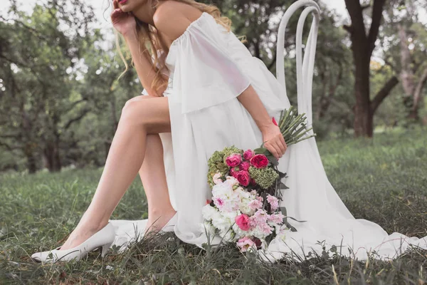 Cropped shot of young bride holding bouquet of flowers and sitting on chair in park — Stock Photo