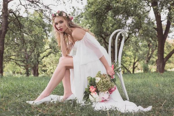 Schöne junge Braut mit Brautstrauß und Blick in die Kamera, während sie auf einem Stuhl im Park sitzt — Stockfoto