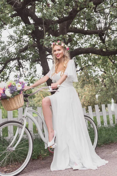 Young blonde bride in wedding dress and floral wreath riding bicycle and smiling at camera — Stock Photo